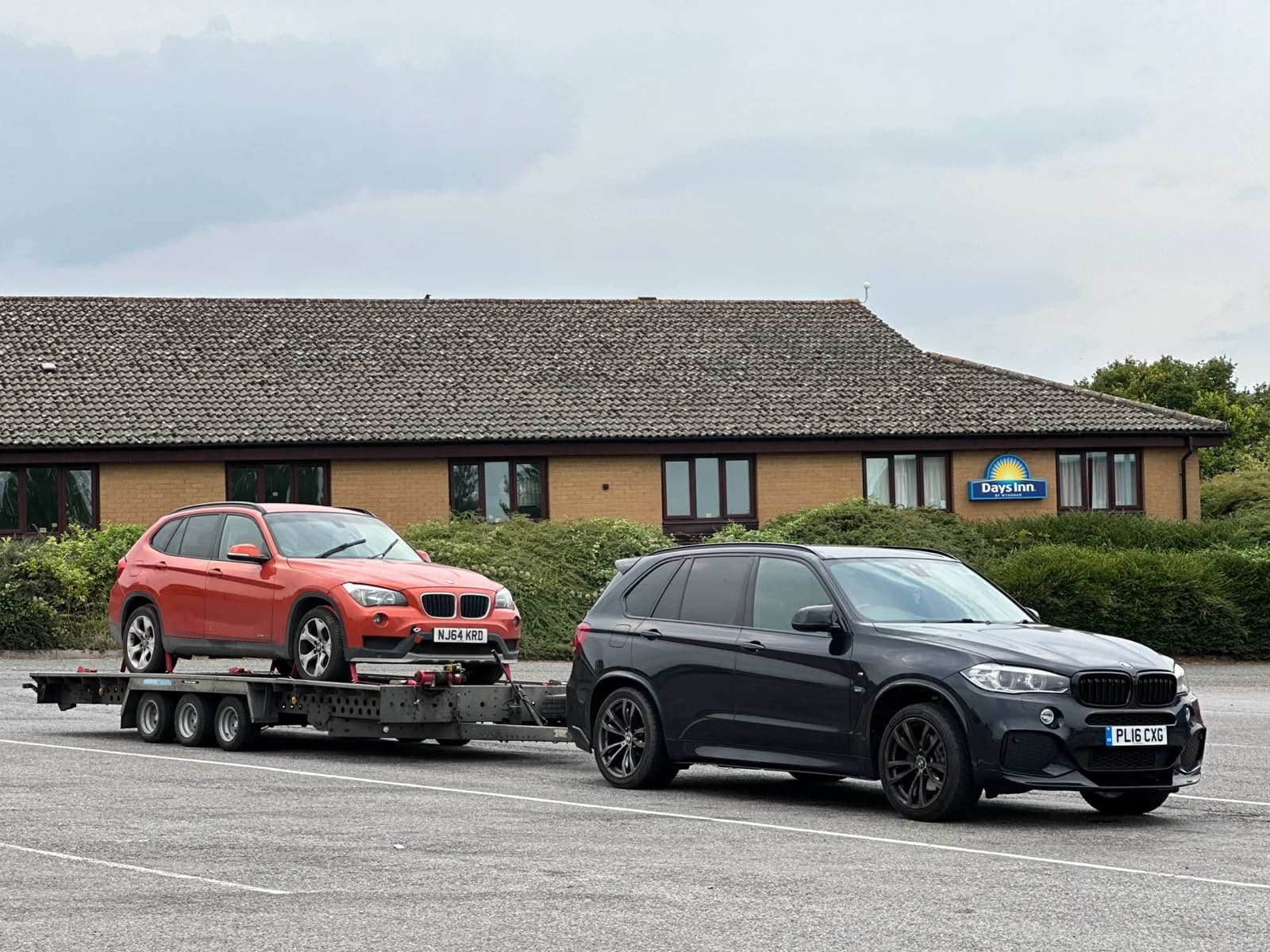 A BMW X5 towing an orange BMW X1 on a trailer