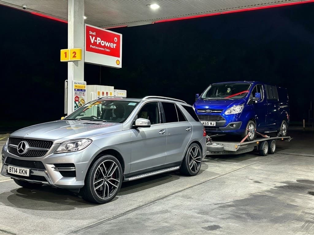 A Mercedes ML towing a blue Ford van on a trailer at night